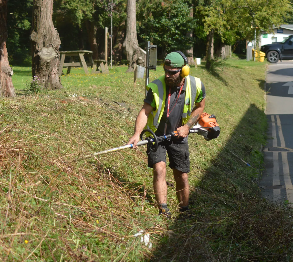 Silver tree services staff use a strimmer to cut back an embankment next to a road